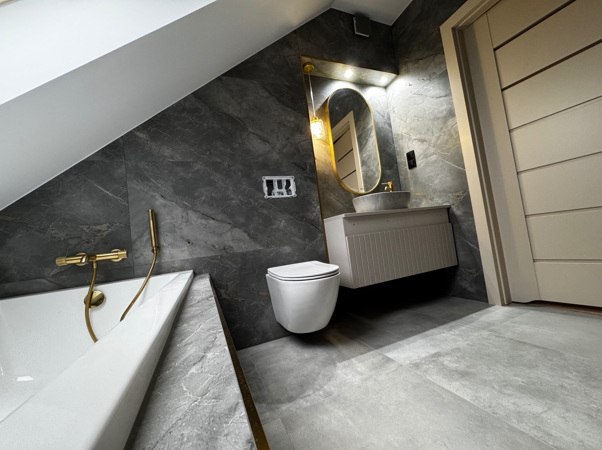 Nowoczesna łazienka na poddaszu z szarymi płytkami imitującymi marmur, złotą armaturą i owalnym lustrem z podświetleniem. Uwagę zwraca skośny sufit.