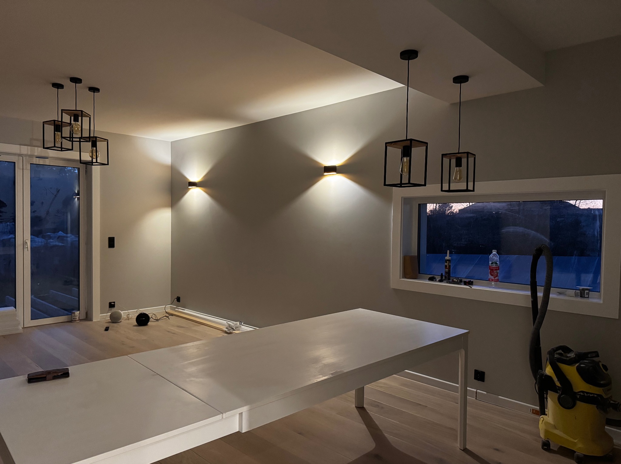 Jasne wnętrze po remoncie w Szczecinku z nowoczesnym oświetleniem: lampy wiszące i ścienne. Widoczny stół, okno i odkurzacz. Ściany w odcieniach szarości.