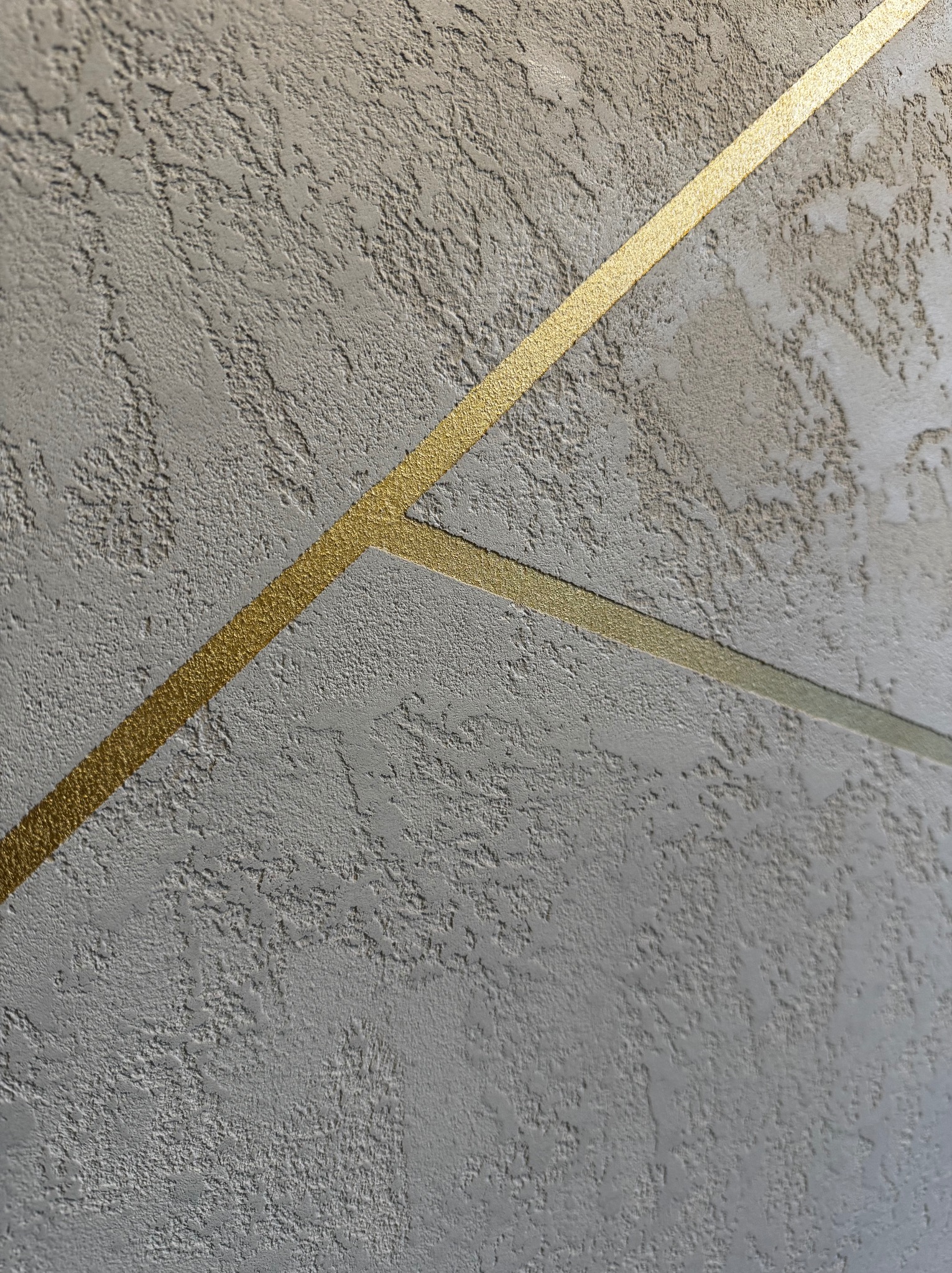 Szara ściana z dekoracyjną fakturą i złotymi, przecinającymi się liniami, tworzącymi geometryczny wzór. Minimalistyczny design.