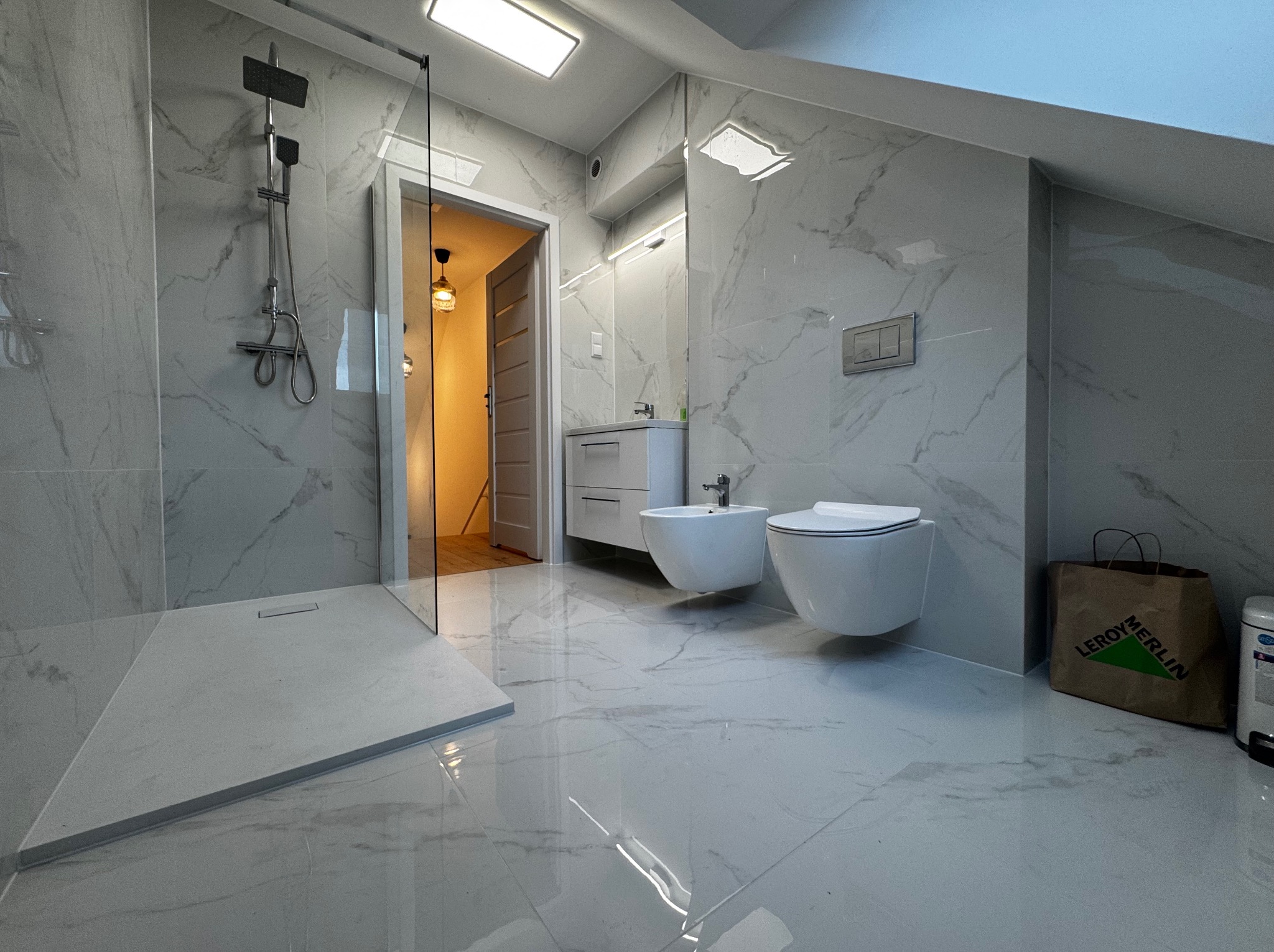 Wykończona łazienka na poddaszu z kabiną prysznicową typu walk-in, wiszącą toaletą i bidetem, wykończona płytkami imitującymi marmur. Widoczna torba z Leroy Merlin.