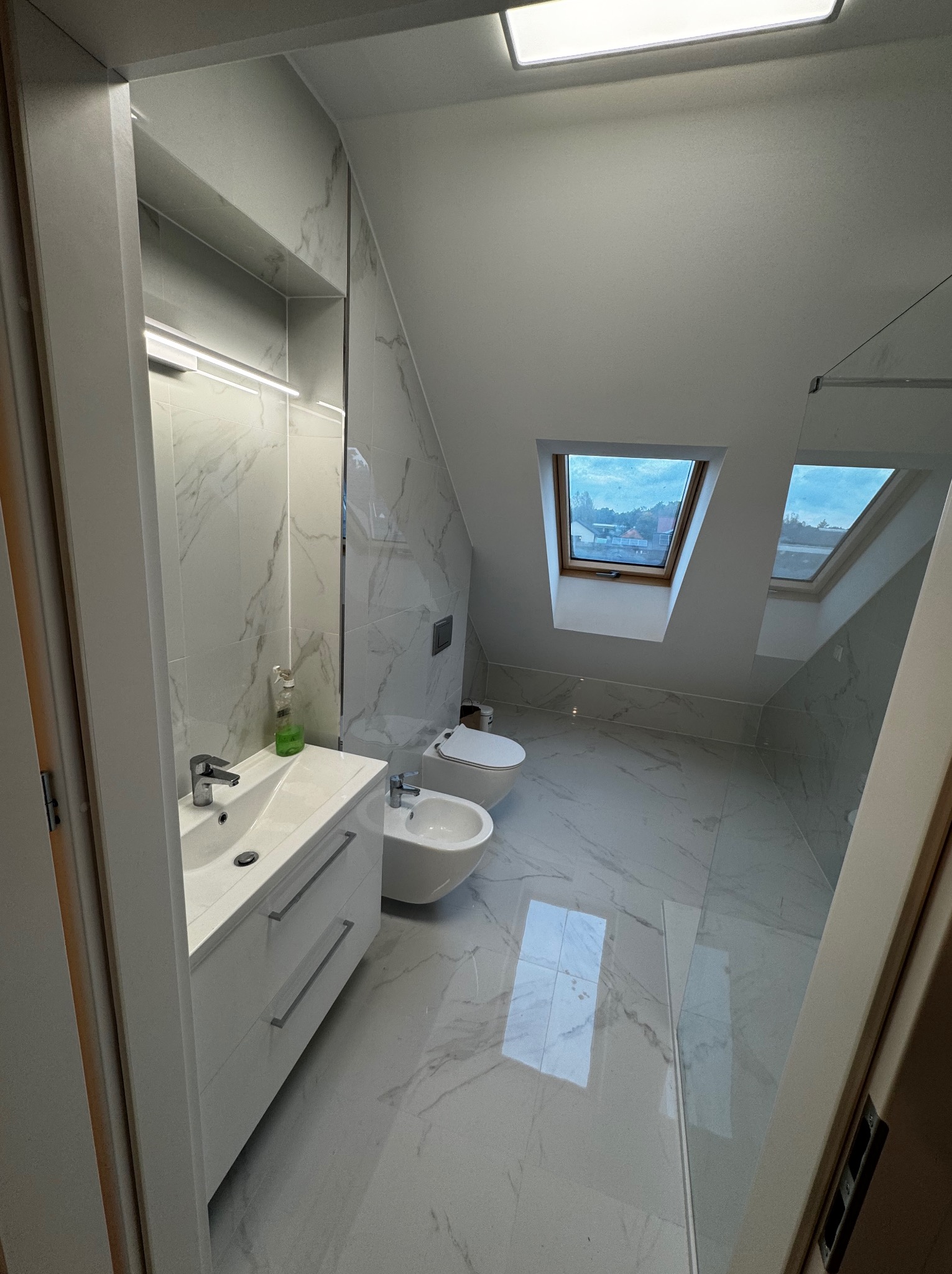 Jasna łazienka na poddaszu z oknami dachowymi, wykończona płytkami imitującymi marmur. Biała szafka z umywalką, bidet i toaleta. Minimalistyczny design.