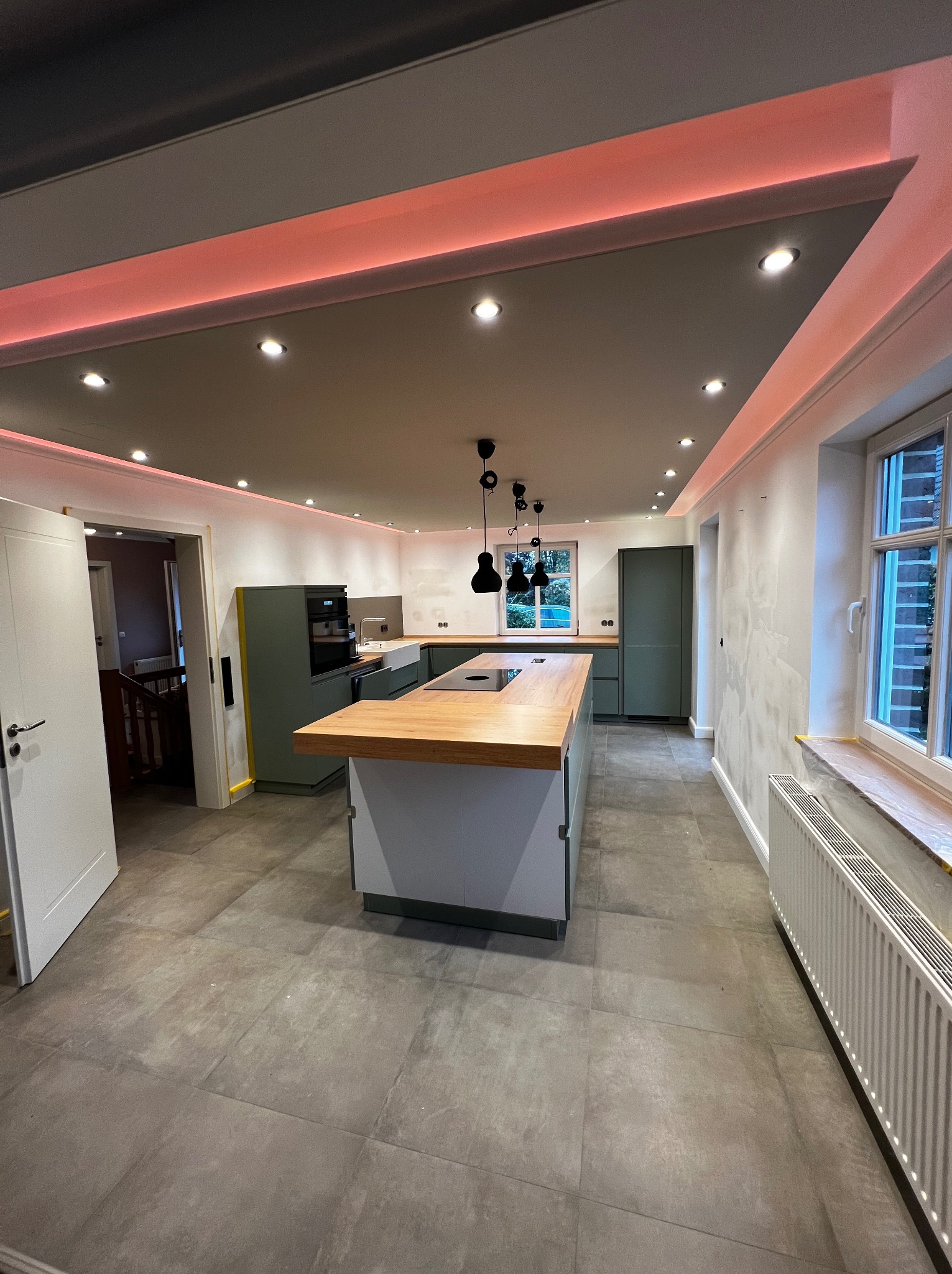 Nowoczesna kuchnia z wyspą, szarymi szafkami i drewnianym blatem. Sufit z oświetleniem LED. Podłoga wyłożona szarymi płytkami. Widoczne okno i grzejnik.