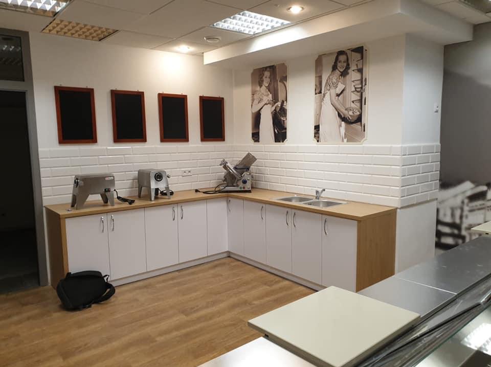 Biała kuchnia z drewnianym blatem, wykończona białymi płytkami imitującymi cegłę, z czarnymi tablicami i czarno-białymi portretami na ścianie. Widoczne sprzęty kuchenne.