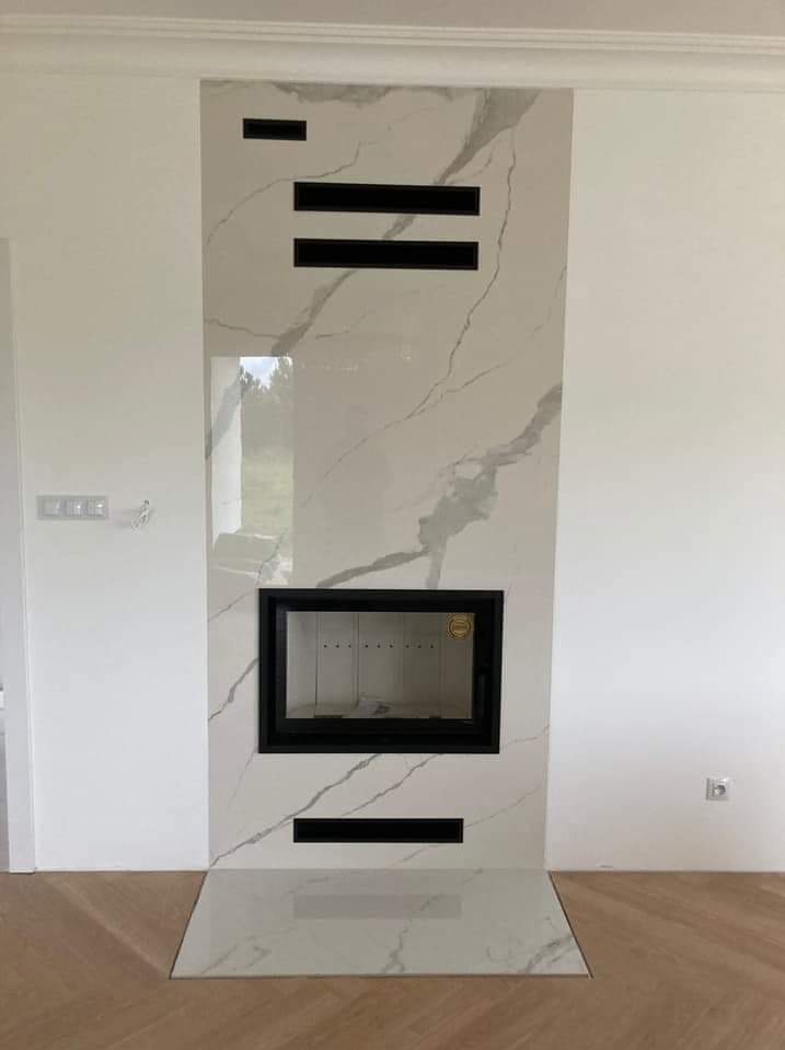 Wykończony kominek z białym marmurem i czarnymi akcentami w nowoczesnym salonie z parkietem w jodełkę. Widoczne gniazdko elektryczne i włącznik światła.