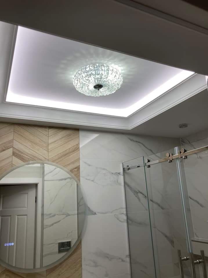 Wykończona łazienka z okrągłym lustrem, marmurowymi płytkami i szklaną kabiną prysznicową. Sufit podwieszany z oświetleniem LED i kryształowym żyrandolem.