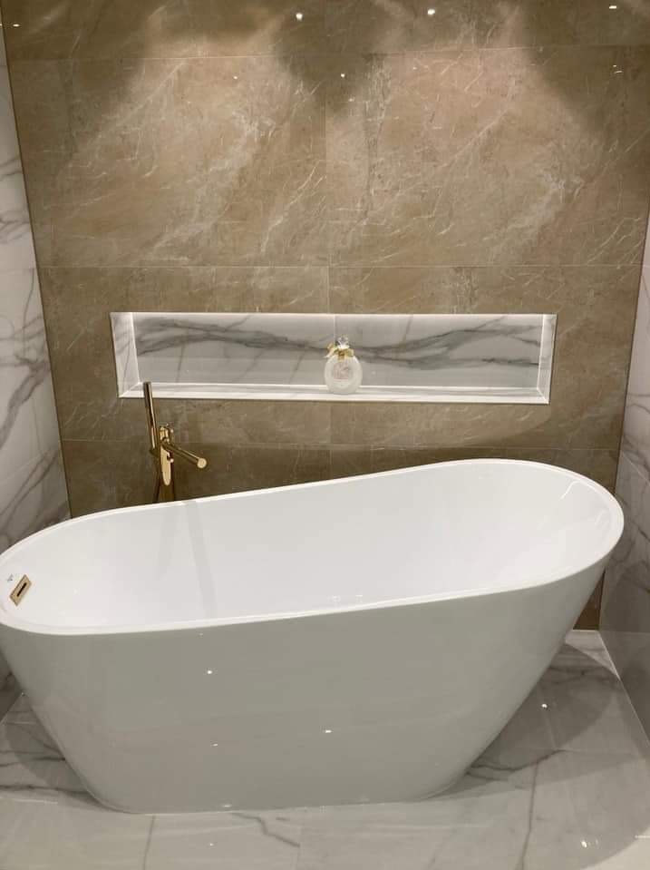 Elegancka łazienka z wolnostojącą wanną, beżowymi płytkami imitującymi marmur i podświetlaną wnęką z dekoracyjnym flakonem. Złota bateria dodaje luksusowego akcentu.