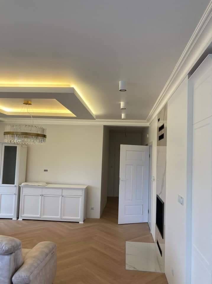 Jasne wnętrze po remoncie: parkiet w jodełkę, białe meble, dekoracyjny sufit z oświetleniem LED i listwy ozdobne. Widok na otwarte drzwi i fragment sofy.