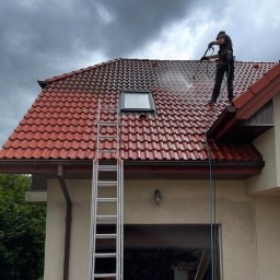 Mazo Wash Krystian Balcerzak - Częściowo umyty dach z czerwonej dachówki, widoczna drabina i osoba w uprzęży czyszcząca dach myjką ciśnieniową. Kontrast czystej i brudnej powierzchni.