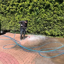 Mazo Wash Krystian Balcerzak - Mężczyzna w ciemnym ubraniu czyści kostkę brukową myjką ciśnieniową na tle zielonego żywopłotu. Widoczne różnice w czystości kostki.