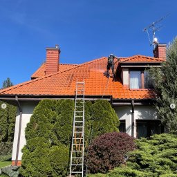 Mazo Wash Krystian Balcerzak - Pracownik w kombinezonie, z drabiny, czyści dach z czerwonej dachówki za pomocą myjki ciśnieniowej. Część dachu umyta, widoczna różnica. Słoneczny dzień.