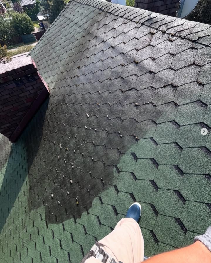 Częściowo umyty dach z gontu bitumicznego, widoczny fragment komina i noga osoby wykonującej prace. Kontrast czystej i zabrudzonej powierzchni.