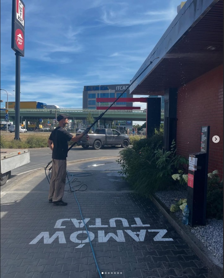 Pracownik w trakcie czyszczenia elewacji budynku KFC/Pizza Hut za pomocą lancy teleskopowej. Widoczne krople wody i napis na kostce brukowej: ZAMÓW TUTAJ.