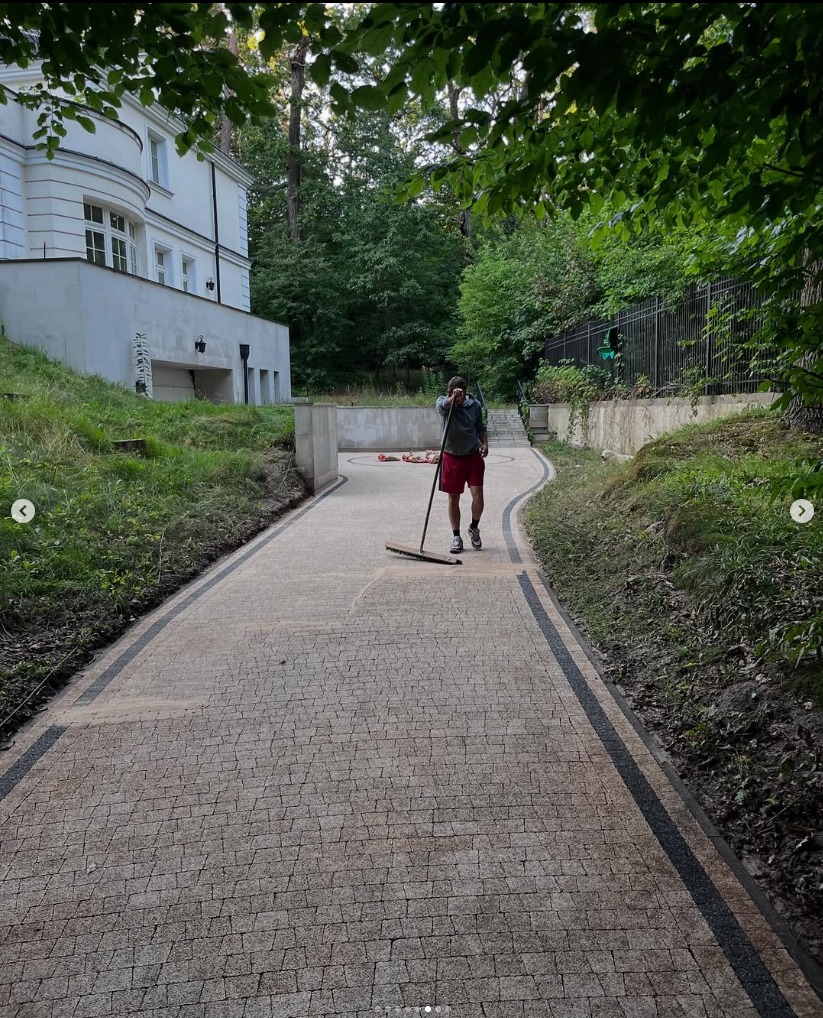 Mężczyzna zamiata kostkę brukową na posesji z białym budynkiem w tle. Zakrzywiona ścieżka, otoczenie zielenią, częściowo zacienione przez drzewa.