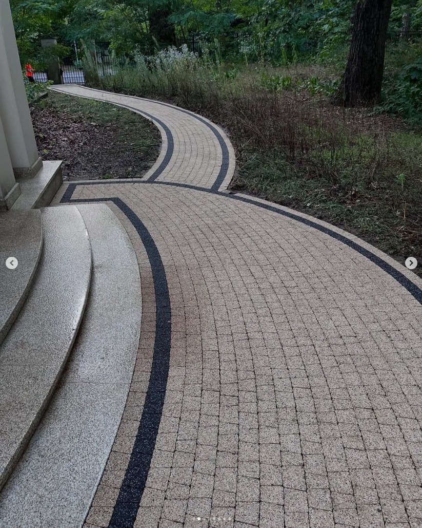 Kaskadowe schody przechodzą w wijący się chodnik z kostki brukowej w odcieniach beżu, obramowany czarnym pasem, prowadzący przez zielony ogród.
