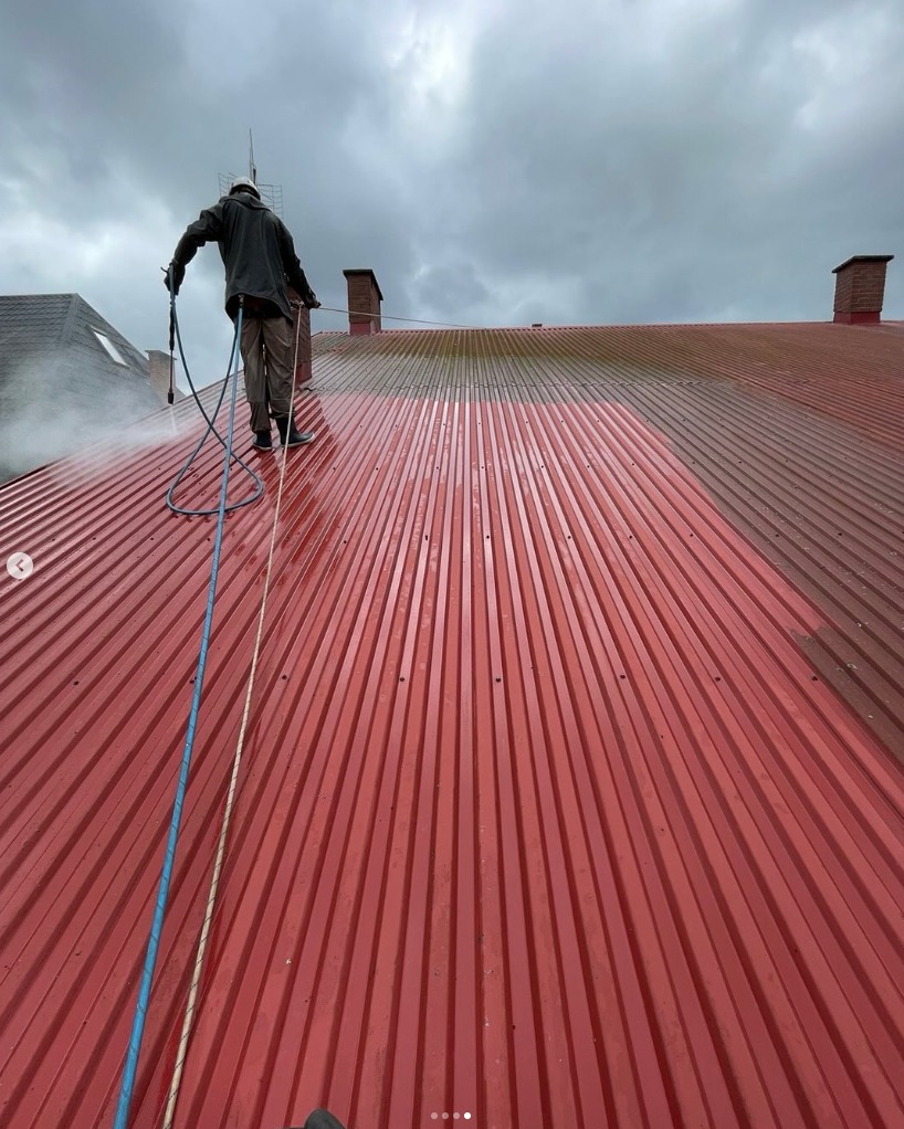 Czerwony dach z blachy trapezowej, częściowo umyty pod ciśnieniem. Widoczna osoba w trakcie pracy z lancą i węże. Pochmurne niebo w tle.