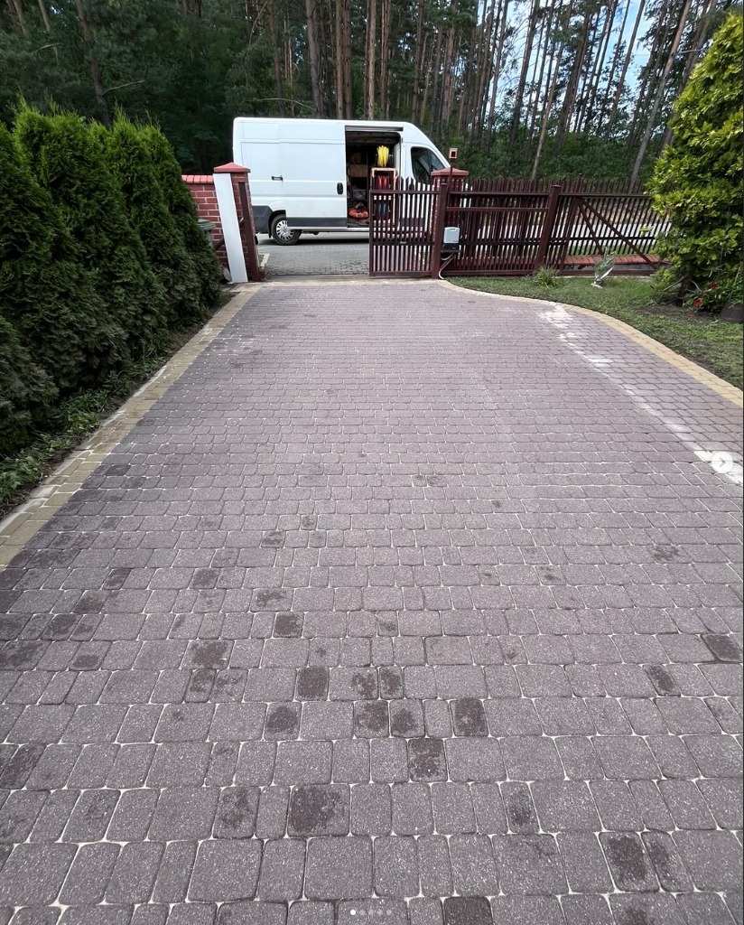 Szara kostka brukowa na podjeździe z widocznymi plamami, brama wjazdowa i biały bus z otwartymi drzwiami w tle, otoczona zielenią.