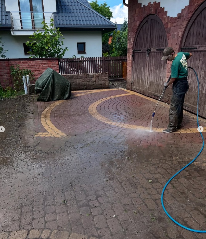 Pracownik w zielonej koszulce czyści kostkę brukową na posesji za pomocą myjki ciśnieniowej. Widoczny efekt czyszczenia i różnica w kolorze kostki.