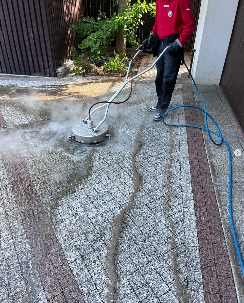Czyszczenie kostki brukowej myjką ciśnieniową z obrotową dyszą. Widoczne ślady po czyszczeniu i para wodna. Osoba w czerwonej bluzie obsługuje urządzenie.