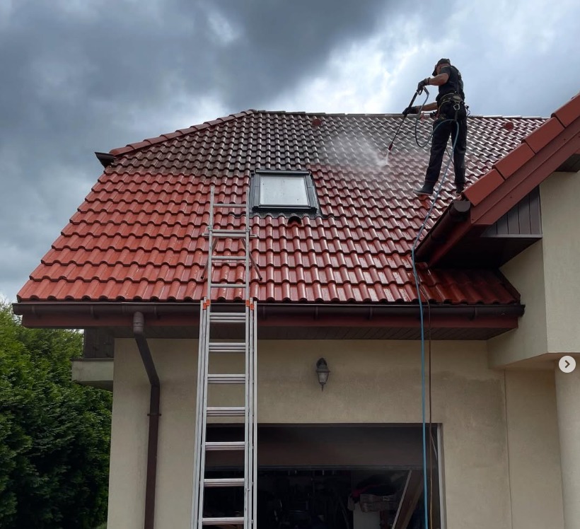 Częściowo umyty dach z czerwonej dachówki, widoczna drabina i osoba w uprzęży czyszcząca dach myjką ciśnieniową. Kontrast czystej i brudnej powierzchni.