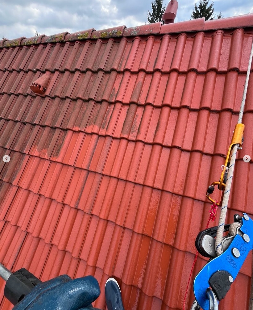 Częściowo umyty, czerwony dach z dachówki karpiówki. Widoczna lina asekuracyjna i fragment narzędzia do czyszczenia. Porównanie stanu przed i po.