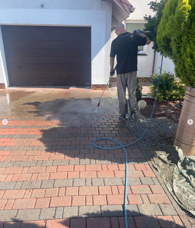 Mężczyzna czyści kostkę brukową przed garażem za pomocą myjki ciśnieniowej. Widoczny efekt czyszczenia i wąż doprowadzający wodę.