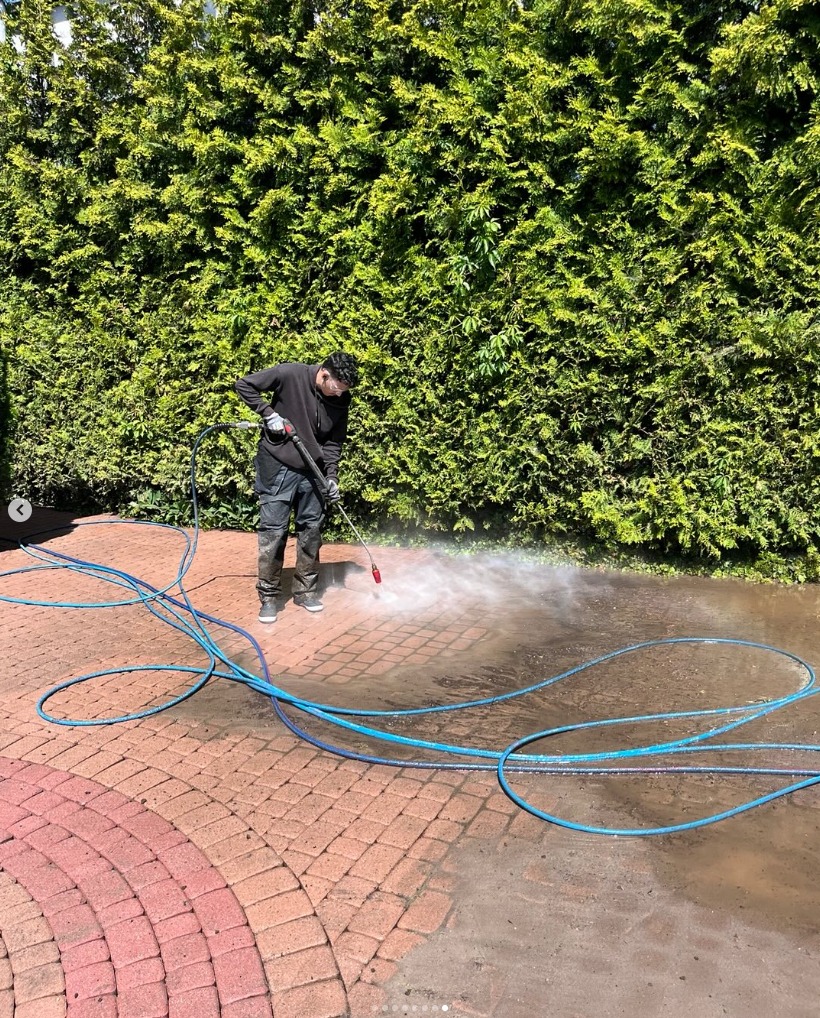 Mężczyzna w ciemnym ubraniu czyści kostkę brukową myjką ciśnieniową na tle zielonego żywopłotu. Widoczne różnice w czystości kostki.