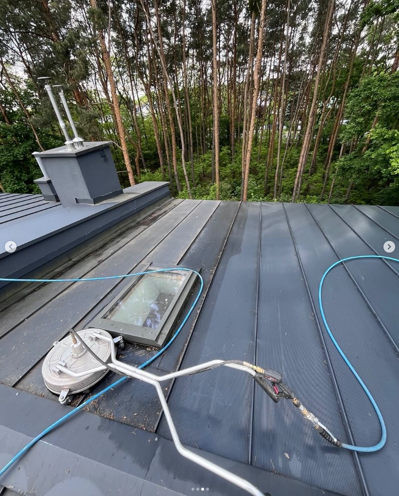 Szary dach z blachy trapezowej, myty specjalistycznym sprzętem ciśnieniowym. Widoczne okno dachowe i fragment lasu w tle. Prace na wysokości.