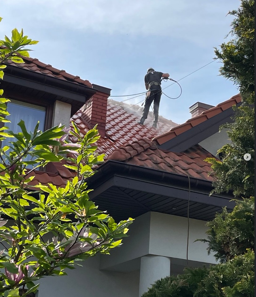 Pracownik w uprzęży czyści dach z czerwonej dachówki za pomocą myjki ciśnieniowej. Widoczne drzewa i fragment domu.