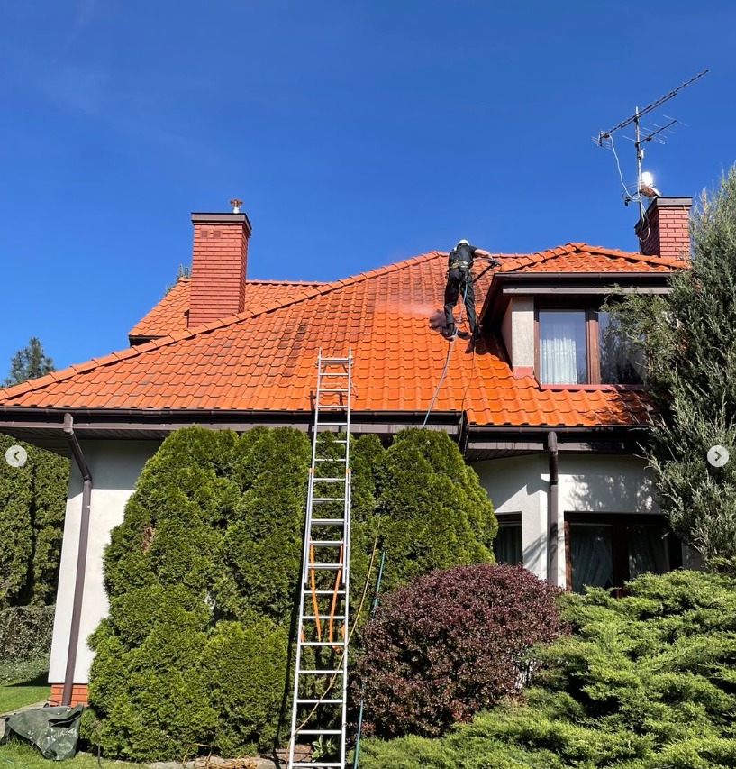 Pracownik w kombinezonie, z drabiny, czyści dach z czerwonej dachówki za pomocą myjki ciśnieniowej. Część dachu umyta, widoczna różnica. Słoneczny dzień.