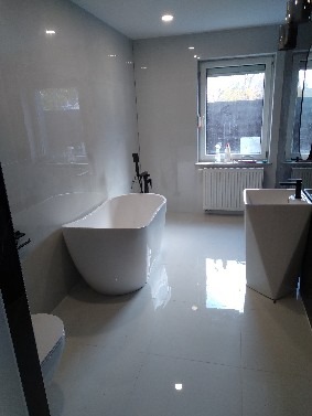 Nowoczesna łazienka z wolnostojącą wanną o obłych kształtach, białą umywalką na postumencie i błyszczącymi, jasnymi płytkami. Minimalistyczny design, widok z perspektywy podłogi.