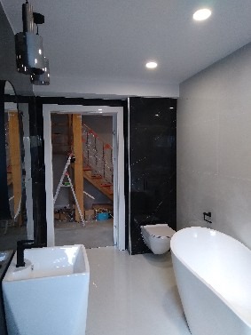 Nowoczesna łazienka w trakcie wykańczania: biała umywalka na postumencie, wanna wolnostojąca, czarne płytki na ścianie, widok na schody przez otwarte drzwi.