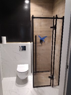 Nowoczesna łazienka z kabiną prysznicową typu walk-in z czarnymi profilami, białą toaletą wiszącą i marmurowymi płytkami na ścianach. Minimalistyczny design.