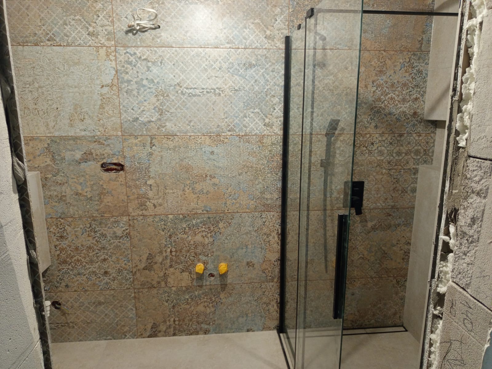 Łazienka w trakcie wykończenia: płytki z wzorem, kabina prysznicowa z czarnymi okuciami, widoczne instalacje i pianka montażowa. Etap prac wykończeniowych.