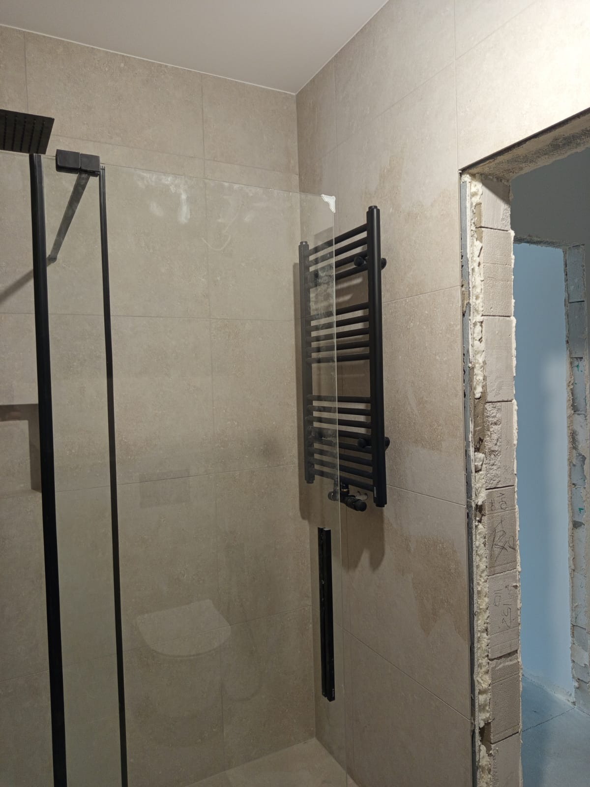 Beżowa łazienka z czarną kabiną prysznicową i grzejnikiem drabinkowym. Widoczne płytki i surowy otwór drzwiowy. Minimalistyczny design.