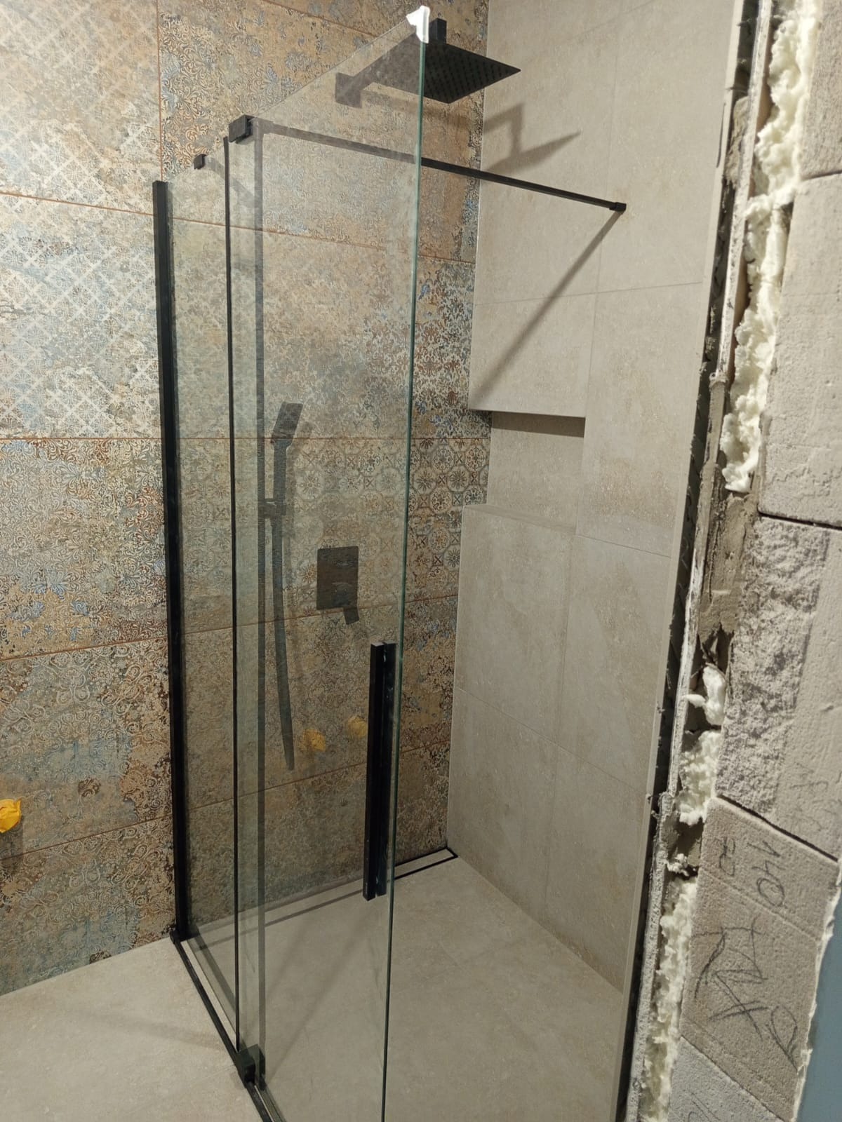 Kabina prysznicowa walk-in z czarnymi profilami na tle płytek w odcieniach beżu i brązu. Widoczne detale wykończenia i fragment ściany w trakcie prac.