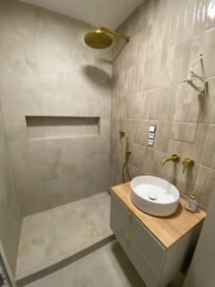 Nowoczesna łazienka z beżowymi płytkami, złotą armaturą i okrągłą umywalką nablatową. W ścianie wnęka. Minimalistyczny design, ciepłe oświetlenie.