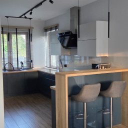 MAR-MEB Mariusz Markowski - Nowoczesna kuchnia z szarymi szafkami, drewnianym blatem i barkiem z hokerami. Oświetlenie punktowe i podłużne lampy wiszące. Parkiet na podłodze.