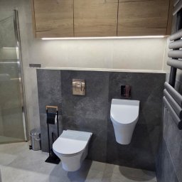 MAR-MEB Mariusz Markowski - Nowoczesna łazienka z podwieszanymi toaletami, szarą płytką i drewnianymi szafkami podświetlonymi LED, minimalistyczny design, widok na kabinę prysznicową.