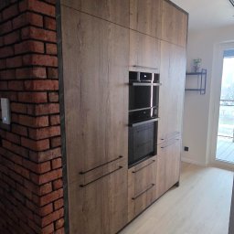 MAR-MEB Mariusz Markowski - Nowoczesna zabudowa kuchenna z forniru drewnopodobnego, wkomponowana w ścianę z cegły. Minimalistyczne uchwyty, piekarnik i mikrofalówka w zabudowie. Oświetlenie szynowe.