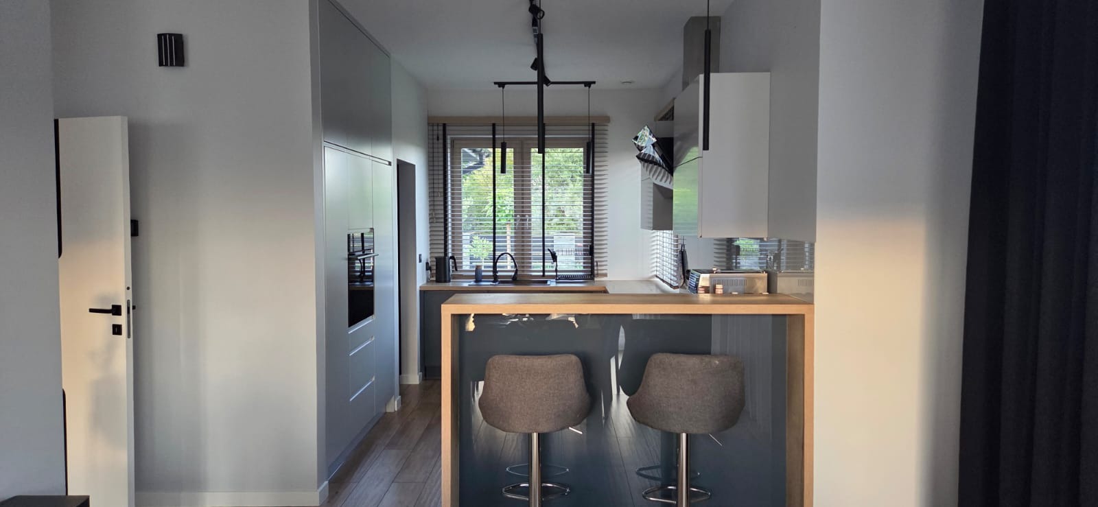 Nowoczesna kuchnia z szarymi szafkami, drewnianym blatem i hokerami. Minimalistyczne oświetlenie i żaluzje w oknie. Widok na wnętrze kuchni z perspektywy baru.