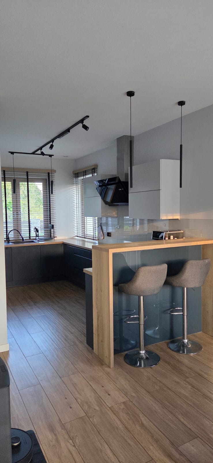 Nowoczesna kuchnia z szarymi szafkami, drewnianym blatem i barkiem z hokerami. Oświetlenie punktowe i podłużne lampy wiszące. Parkiet na podłodze.