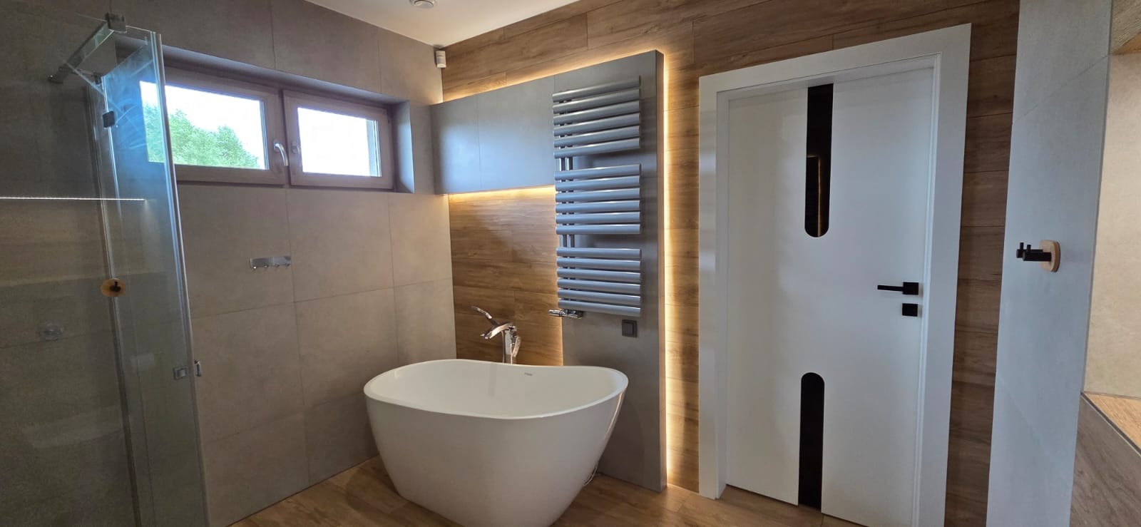 Nowoczesna łazienka z wolnostojącą wanną, grzejnikiem drabinkowym podświetlonym LED i designerskimi drzwiami z czarnymi akcentami. Uwagę zwraca drewnopodobna okładzina.