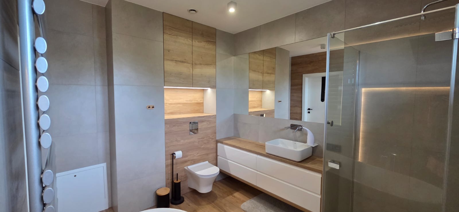 Nowoczesna łazienka z drewnianymi akcentami: szafki, blat pod umywalką, płytki. Kabina prysznicowa ze szklanymi drzwiami, podświetlane lustro i minimalistyczna toaleta.