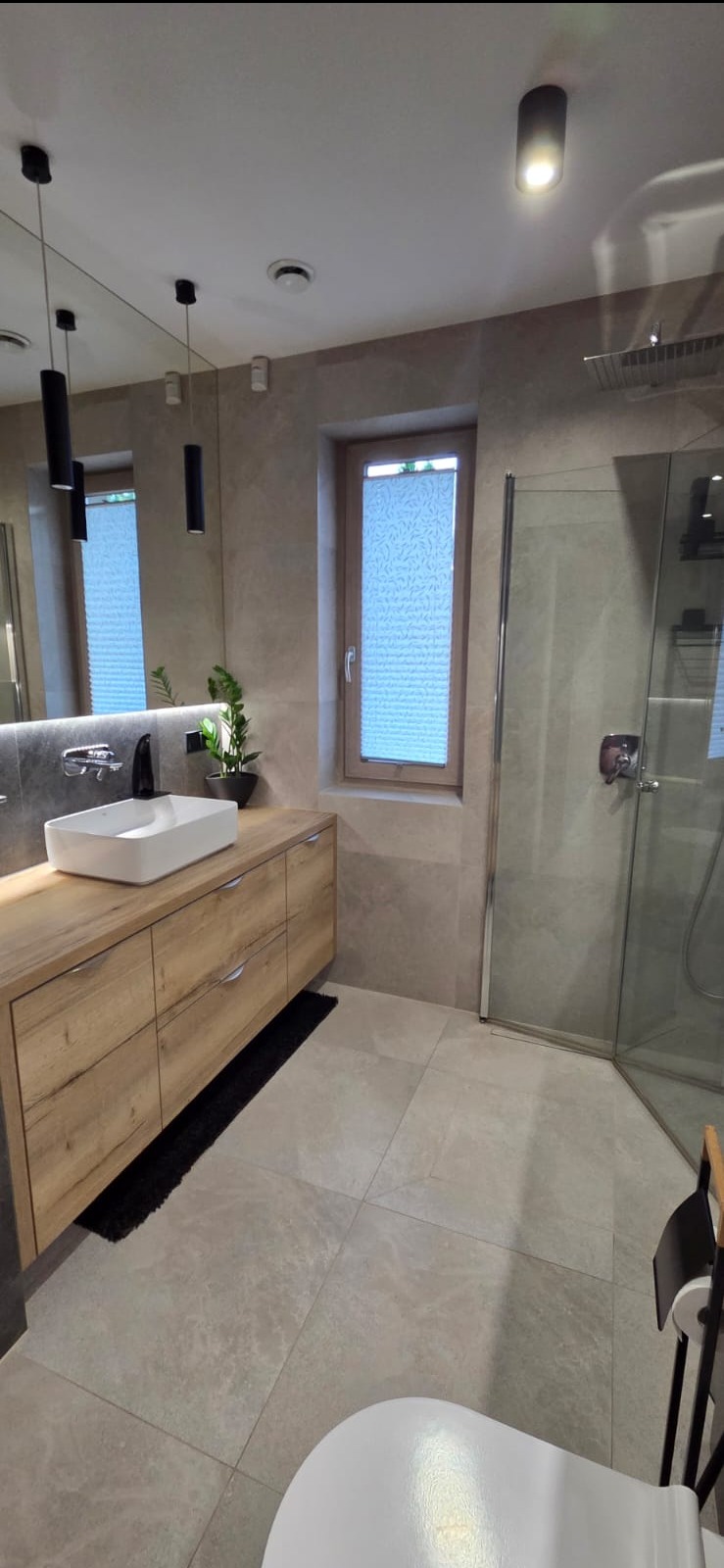 Nowoczesna łazienka z drewnianą szafką pod umywalką, kabiną prysznicową i oknem z roletą. Stonowana kolorystyka, minimalistyczne oświetlenie.