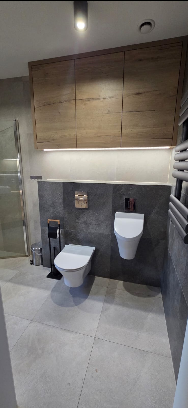 Nowoczesna łazienka z podwieszanymi toaletami, szarą płytką i drewnianymi szafkami podświetlonymi LED, minimalistyczny design, widok na kabinę prysznicową.