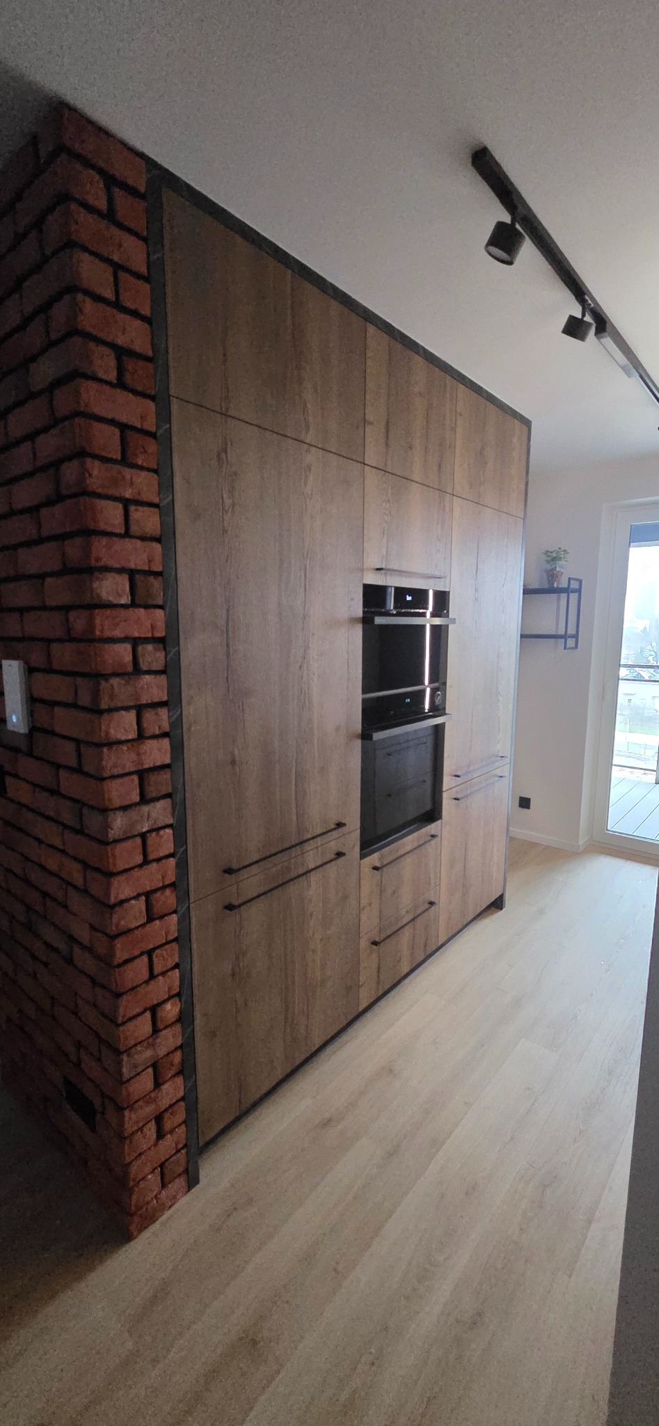 Nowoczesna zabudowa kuchenna z forniru drewnopodobnego, wkomponowana w ścianę z cegły. Minimalistyczne uchwyty, piekarnik i mikrofalówka w zabudowie. Oświetlenie szynowe.