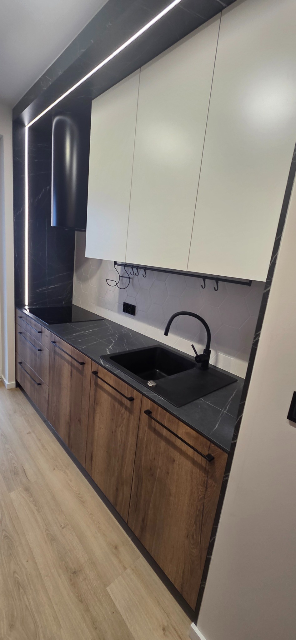 Nowoczesna kuchnia z drewnianymi szafkami, czarnym blatem i armaturą. Białe górne szafki i heksagonalne płytki dodają elegancji. Oświetlenie LED podkreśla marmurowy wzór.