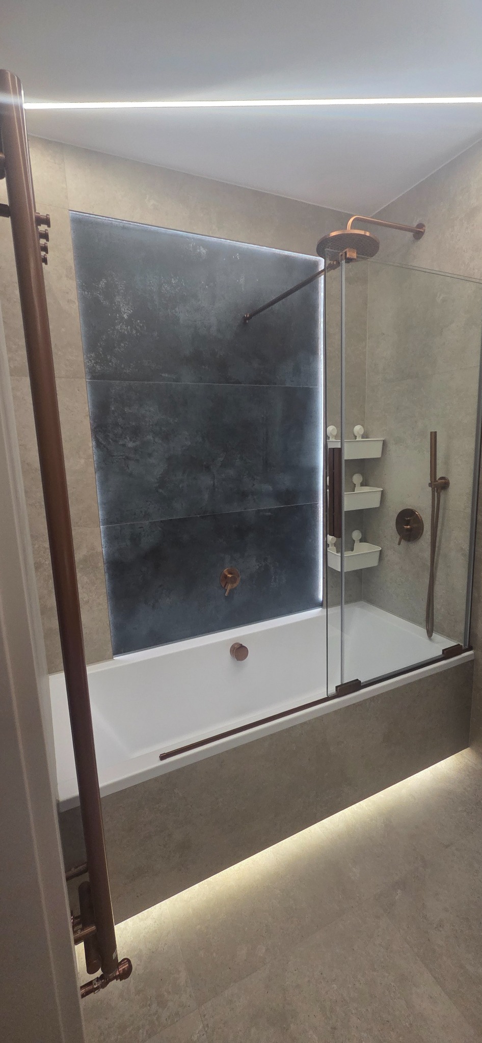 Nowoczesna łazienka z wanną i szklanym parawanem, akcenty miedziane, podświetlenie LED pod wanną i za płytkami ściennymi. Minimalistyczny design, szaro-biała kolorystyka.