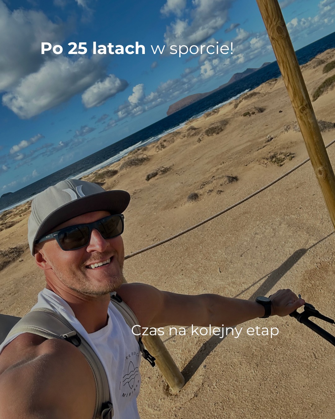 Uśmiechnięty mężczyzna w okularach przeciwsłonecznych i czapce, robiący selfie na tle plaży z widokiem na morze i góry. Napis: Po 25 latach w sporcie! Czas na kolejny etap.