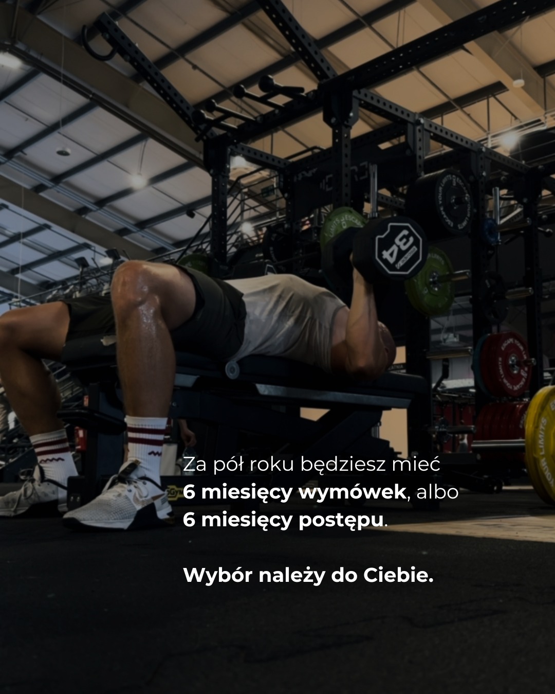 Mężczyzna ćwiczy wyciskanie hantli na ławce w siłowni, widoczne obciążenia i konstrukcja do ćwiczeń. Na dole motywujący tekst o wyborze postępu.